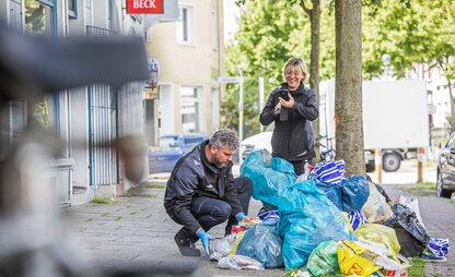 Eine Person kniet vor einem Müllhaufen, der aus mehreren Müllsäcken und Tüten besteht, während eine zweite Person hinter den illegalen Müllablagerungen steht und ein Foto von der Situation macht. | © Die Bremer Stadtreinigung