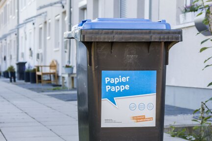 Eine Papiertonne mit blauem Deckel, die vor einer Hausreihe steht. Auf der Tonne steht " Papier, Pappe".