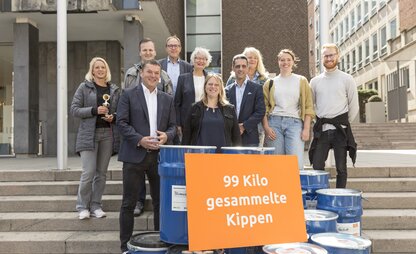 Eine Gruppe steht vor blauen Tonnen, auf dem ein Schild mit der Information steht, dass 99 Kilo Zigarettenkippen gesammelt wurden. | © Die Bremer Stadtreinigung