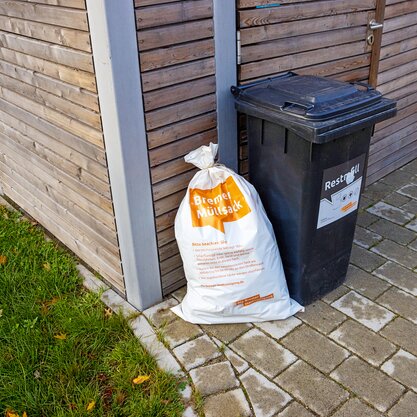 Eine schwarze Tonne mit braunem Deckel auf der "Restmüll" steht. Davor ist ein weißer Sack mit der Aufschrift "Bremer Müllsack". | © Die Bremer Stadtreinigung