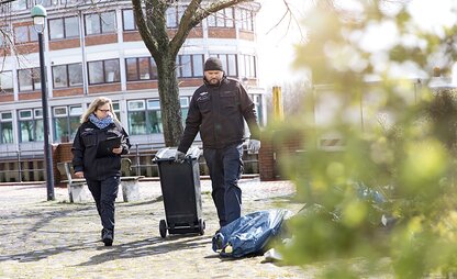 Zwei Mitarbeitende des Kontrolldienstes prüfen illegale Müllablagerungen auf der Straße. Eine Person zieht eine Tonne hinter sich her. | © Die Bremer Stadtreinigung