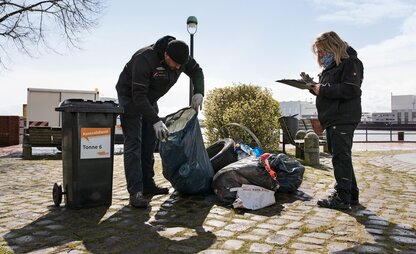 Zwei Mitarbeitende des Kontrolldienstes untersuchen illegale Müllablagerungen. Die linke Person schaut in eine blaue Mülltüte hinein, während die rechte Person Notizen auf einem Klemmbrett macht. | © Die Bremer Stadtreinigung