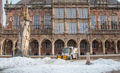 Ein Räumungsfahrzeug des Winterdiensts räumt den Schnee vor dem Bremer Rathaus beiseite. Im Vordergrund ist die Rolandstatue zu sehen. | © Die Bremer Stadtreinigung