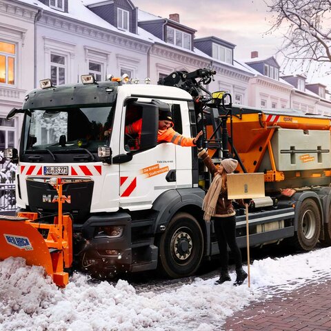 Schneeräumfahrzeug der Bremer Stadtreinigung mit Schneepflug auf verschneiter Straße neben einer Personen mit Schneeschieber, der Fahrer und die Person klatschen ab | © Die Bremer Stadtreinigung