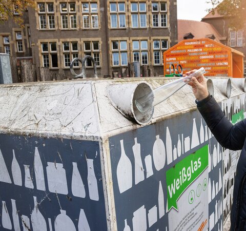 Eine Person wirft eine durchsichtige Glasflasche in die Öffnung eines Containers. Auf dem Container steht Weißglas. | © Die Bremer Stadtreinigung