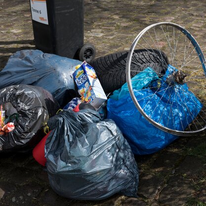 Vor einer Mülltonne sind illegale Müllablagerungen zu sehen: Mehrere Mülltüten, auf der eine Eisteetüte und die Speichen eines Fahrrads liegen. | © Die Bremer Stadtreinigung