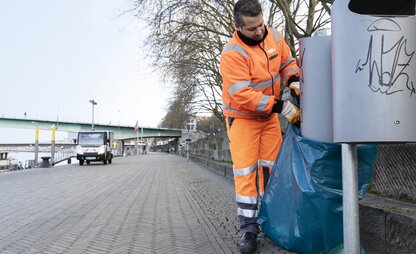 Ein Mitarbeiter von DBS leert an der Bremer Schlachte zwei öffentliche Abfallbehälter. Im Hintergrund ist das Entsorgungsfahrzeug an der Promenade zu sehen. | © Die Bremer Stadtreinigung