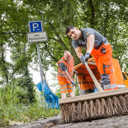 Straßenreinigung mit Besen | © Die Bremer Stadtreinigung Zwei Mitarbeitende von DBS fegen mit einem Besen die Straße. In der Bildmitte lehnt ein Blauer Müllsack an einem Straßenschild, das auf eine Parkfläche hinweist. | © Die Bremer Stadtreinigung
