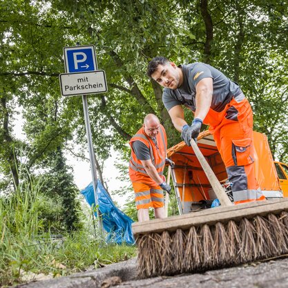 Straßenreinigung mit Besen | © Die Bremer Stadtreinigung Zwei Mitarbeitende von DBS fegen mit einem Besen die Straße. In der Bildmitte lehnt ein Blauer Müllsack an einem Straßenschild, das auf eine Parkfläche hinweist. | © Die Bremer Stadtreinigung